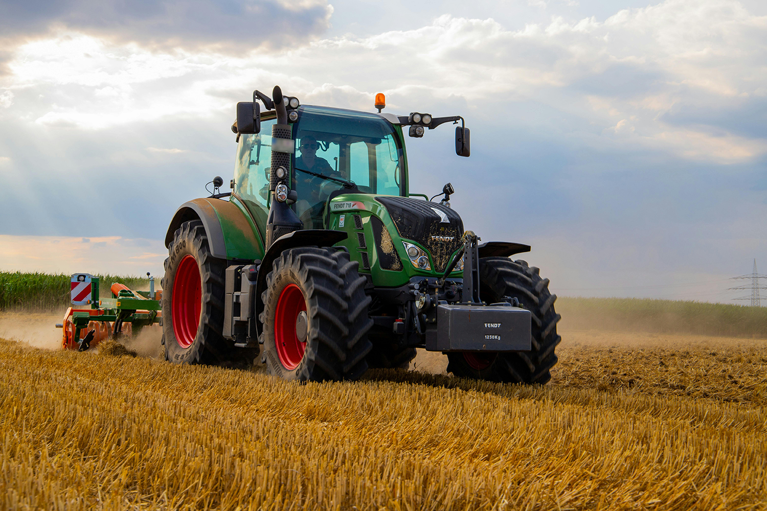 A green and black tractor plowing a field of harvested crops with a cloudy sky in the background.